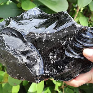 以下が含まれることがあります： 黒曜石の大きな、不規則な形の塊。表面は光沢があり、光を反射し、ひび割れや質感が目に見えます。石は緑の葉を背景に保持されています。