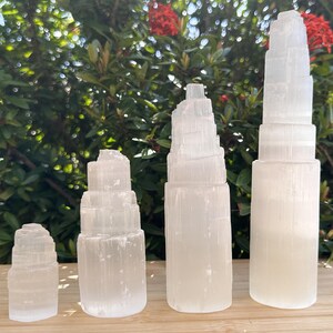 May include: Four white selenite crystal towers of varying heights arranged in a row. Each tower features a cylindrical base that transitions into a stepped, layered structure at the top, showcasing a translucent, fibrous, and crystalline texture.
