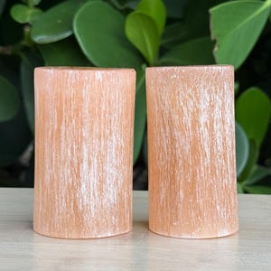May include: Two cylindrical selenite towers with a soft peach-orange hue and a fibrous, translucent texture. Each piece features vertical white striations running along the length of the stone, creating a natural, shimmering appearance.