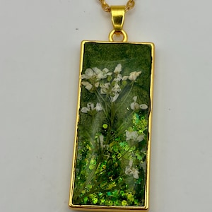 May include: A rectangular pendant necklace featuring a gold-toned metal frame. The interior contains a dark green background with embedded white pressed flowers and bright green glitter particles. The pendant hangs from a matching gold-toned chain.