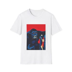 May include: White t-shirt featuring a graphic of a giant gorilla with red eyes and a woman in a red dress. The gorilla is dark blue, set against a red background. The woman is blonde and appears to be in distress.