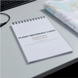 May include: A white spiral-bound notebook with the title "FLIGHT REFERENCE CARDS" and the subheadings "Operational Checklists" and "Emergency Checklists". A map is partially visible in the background. The notebook is on a light grey surface.