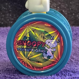 May include: A Snoopy and the Peanuts Gang yo-yo toy. The yo-yo is teal with a holographic image of Snoopy and the Peanuts Gang. A white Snoopy figure is on top of the yo-yo, with his arm raised.