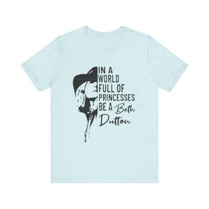 May include: Light blue t-shirt with a black graphic of a woman's silhouette and the text "IN A WORLD FULL OF PRINCESSES BE A Beth Dutton". The shirt is made of a soft material.