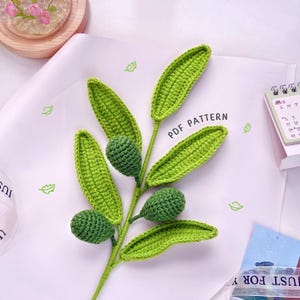 Può includere: Un ramo di ulivo all'uncinetto fatto a mano, con foglie e olive verdi. Il ramo è realizzato in filato e ha un aspetto strutturato. Il testo "PDF PATTERN" è visibile sulla carta. Un progetto artigianale.