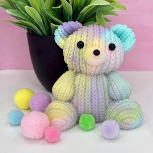 May include: A pastel-colored knitted teddy bear with black button eyes and a variety of colorful pom-poms. The bear is primarily yellow, purple, and blue. A black pot with green leaves is in the background.