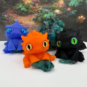 May include: Three small, plush dragon figurines in blue, orange, and black. Each dragon has large, round eyes with a contrasting iris color. The orange dragon is in the center, with a blue dragon on the left and a black dragon on the right. Each dragon is resting on a piece of green crystal.