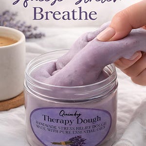 May include: A jar of purple therapy dough with the text "Squeeze. Stretch. Breathe." and "Quinky Therapy Dough." The dough is being stretched by a hand. The jar is on a white surface, with a cup of coffee and lavender sprigs in the background.