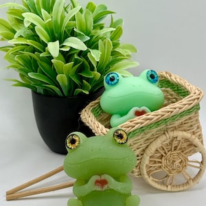 May include: Two green frog figurines with large, expressive eyes, one holding a heart. A small woven cart with a frog inside and a potted green plant are also present. The frogs have a charming, whimsical design.