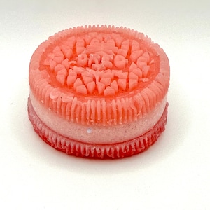 May include: A pink and white decorative item resembling a cookie. The top layer is a textured pink with the word "OREO" embossed. The middle layer is white, and the bottom layer is a darker pink with a ridged edge. The item is on a white background.