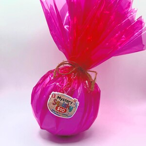 May include: A bright pink, wrapped gift with a decorative top. The gift is tied with twine and features a sticker that reads "Mystery Squishy $10". The gift is a surprise.