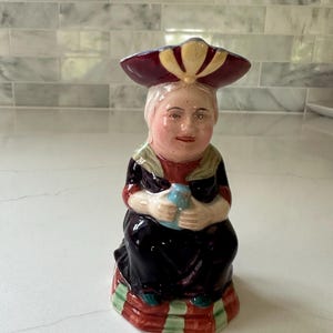 May include: A ceramic figurine of a person wearing a dark purple dress, a light green shawl, and a wide-brimmed maroon hat with cream accents. The figure holds a small light blue object in their hands and sits on a base with red and green stripes.