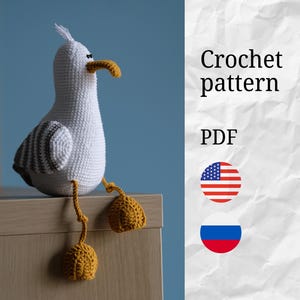 Op de afbeelding: Een handgemaakt gehaakt zeemeeuw speelgoed in wit, grijs en geel. De zeemeeuw heeft een witte body, grijze vleugels en een gele snavel en poten. De tekst "Crochet pattern PDF" wordt aan de rechterkant van de afbeelding weergegeven.