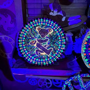 May include: A round, black, glowing art piece featuring a dancing bear design. The bear is surrounded by a colorful dotted pattern in shades of blue, green, and orange. The artwork is displayed in a dimly lit room with other glowing objects.