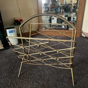 Puede incluir: Un revistero de metal dorado con asa. El revistero tiene un diseño geométrico con una serie de líneas que se cruzan y pequeños detalles de bolas. Se apoya en cuatro patas delgadas.