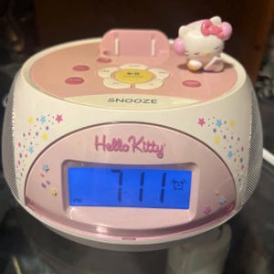 May include: A pink and white Hello Kitty alarm clock with a digital display showing the time as 7:11 PM. The clock features a Hello Kitty figurine on top and the word "SNOOZE". The clock is decorated with star and dot patterns.