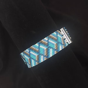 May include: A woven bead bracelet featuring a repeating geometric pattern in shades of light blue, turquoise, white, and metallic bronze. The bracelet is secured with a polished silver-toned metal slide clasp that connects the two ends together.