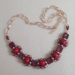 May include: A necklace featuring a rose gold-toned chain and a central arrangement of red marbled spherical beads, square beads with etched starburst patterns, and small clear crystal accents, all connected with intricate beadwork and metal links.