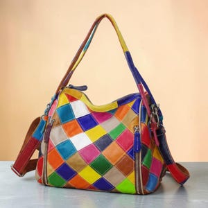 Puede incluir: Un bolso de mano multicolor con un diseño de patchwork de piezas de cuero en forma de rombo en tonos azul, amarillo, naranja, verde, rojo y blanco. El bolso incluye dos asas superiores, una correa de hombro desmontable y cremalleras plateadas.