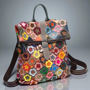 Può includere: Zaino colorato con un design patchwork, adornato da numerose applicazioni di fiori in tessuto. La borsa è dotata di una maniglia superiore, spallacci regolabili e una chiusura con fibbia. La tavolozza dei colori comprende tonalità di arancione, blu, rosa e verde.
