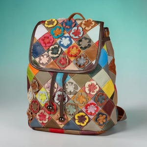 Puede incluir: Una mochila multicolor con un diseño de patchwork en forma de rombos. El exterior está adornado con numerosas pequeñas aplicaciones de flores de cuero superpuestas en varios colores, incluyendo rojo, amarillo, azul y blanco. Tiene cierre de cordón.