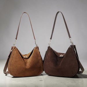 Puede incluir: Dos bolsos de hombro estilo hobo hechos de ante, uno en color marrón claro y otro en marrón chocolate oscuro. Cada bolso cuenta con un asa superior, un cierre giratorio plateado en el centro y detalles de costuras decorativas en los bordes.