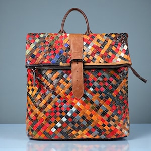 Op de afbeelding: Een kleurrijke, geweven leren rugzak met een handvat aan de bovenkant en een bruine leren riem. De tas heeft een ritssluiting en een verscheidenheid aan kleuren, waaronder rood, oranje, blauw en groen. De tas is ongeveer 30 cm breed en 36 cm hoog.