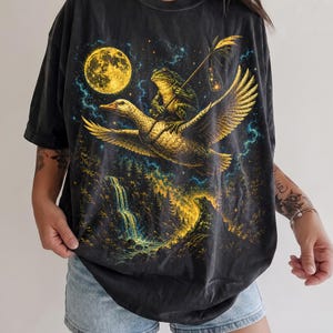 Pode incluir: Uma t-shirt preta de manga curta com uma estampa gráfica de um sapo montado num ganso voador contra um céu noturno com lua cheia. A cena inclui uma floresta, uma cascata e uma pequena lanterna segurada pelo sapo, em tons amarelos e azuis.