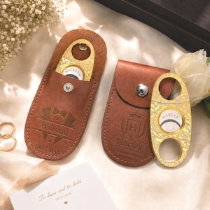 May include: Two brown leather cigar cutter cases with silver snap closures. One case is engraved with a tuxedo design and the name William, the other with a crest, the name Harley, and the date 08.10.2026. Two gold-toned metal cigar cutters are included.