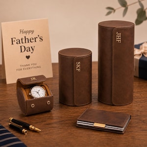 Puede incluir: Un juego de regalo para el Día del Padre con un reloj, estuches de viaje para relojes, una tarjeta y una cartera. El reloj está en un estuche de cuero marrón. La tarjeta dice "Feliz Día del Padre".