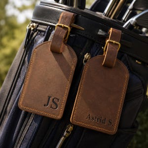 May include: Two rectangular brown leather luggage tags with rounded tops and brass buckles. One tag is embossed with the initials JS, and the other is embossed with the name Astrid S. The tags are attached to a dark navy blue golf bag.