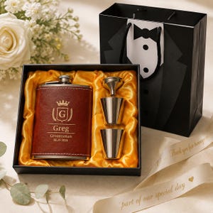 Op de afbeelding: Een bruine, met leer beklede metalen heupfles gegraveerd met een kroon, de letter G, de naam Greg, Groomsman en 08.29.2026, in een zwarte geschenkdoos met gele satijnen voering, twee zilveren borrelglaasjes, een trechter en een smokingtas.
