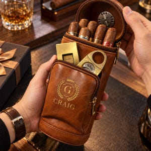 May include: A brown leather cigar case featuring a zippered front pocket with a gold-toned crest logo and the name CRAIG embossed on the front. The case holds several cigars, a gold-toned lighter, and a gold-toned cigar cutter inside the top compartment.