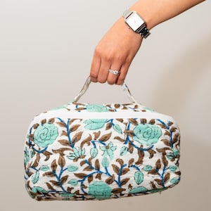 Puede incluir: Un neceser acolchado blanco con un estampado floral en tonos turquesa, marrón y azul. El neceser tiene cierre de cremallera y un asa. El neceser está sostenido por una persona.