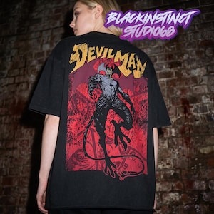 Peut inclure: Un t-shirt noir avec une grande impression graphique au dos. Le motif présente le texte DEVILMAN dans une police jaune vieillie au-dessus d'une créature sombre et musclée avec des ailes et une longue queue, sur un fond rouge vif.