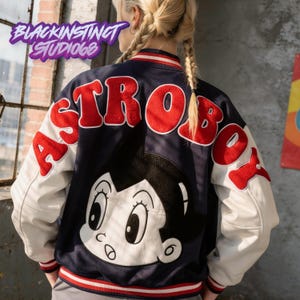 May include: Navy blue and white Astro Boy varsity jacket with red lettering and a large Astro Boy graphic on the back. The jacket has white sleeves and the text "BLACKINSTINCT STUDIOS68" in purple.