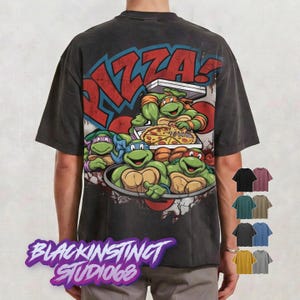 May include: Dark gray t-shirt featuring a graphic of the Teenage Mutant Ninja Turtles with the word "PIZZA!" in red and blue. The shirt also has the text "BLACKINSTINCT STUDIO68" at the bottom. Several color options are displayed.
