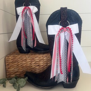 May include: A pair of black cowboy boots with decorative floral patterns. Each boot is adorned with a white bow and cascading ribbons in red gingham, white lace, and red pom-pom trim. The boots are displayed on a woven basket.
