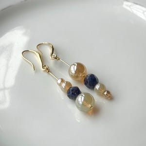 May include: A pair of gold-toned dangle earrings. Each earring features a series of beads in shades of gold, blue, and clear, strung on a delicate gold wire. The earrings are displayed on a white plate.