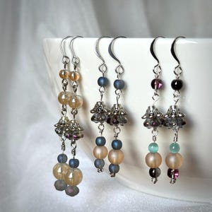 May include: Three pairs of dangle earrings with silver-tone hooks. Each earring features a combination of beads in shades of peach, blue, and purple, along with decorative silver-tone bell-shaped charms. The earrings are displayed against a white background.