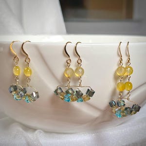 May include: Three pairs of gold-toned dangle earrings. Each earring features yellow beads and a cluster of blue, yellow, and clear crystals. The earrings are displayed in a white bowl.