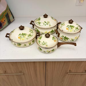 May include: A set of four cream-colored enamelware pots and pans with lids. Each piece features a floral design with green leaves and yellow flowers. The handles and knobs are brown.
