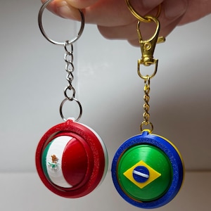 Puede incluir: Dos llaveros con diseños circulares que representan banderas. Uno presenta la bandera mexicana con rojo, blanco y verde. El otro muestra la bandera brasileña con verde, amarillo, azul y blanco. Ambos tienen llaveros de metal.
