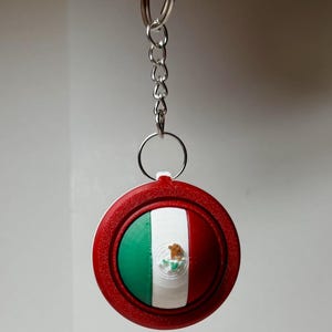 May include: A round keychain featuring the Mexican flag. The keychain has a red border and a central design with green, white, and red stripes, and a coat of arms. The keychain is attached to a silver chain and ring.