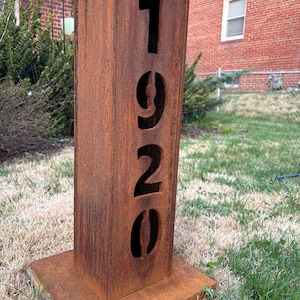 May include: A weathered, rectangular metal address post with the numbers "1920" cut out. The post is a rusty brown color and sits on a matching square base, placed in a grassy yard.