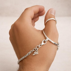 May include: A silver bracelet with a ring attached to a chain. The bracelet has a lobster clasp and a small charm. The ring is on the thumb. The bracelet is made of a chain link design.