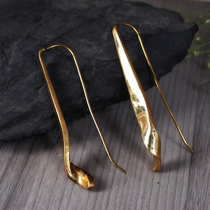 May include: A pair of gold-coloured earrings. The earrings have a long, curved design with a textured, leaf-like shape at the bottom. The earrings are displayed on a dark, textured surface.
