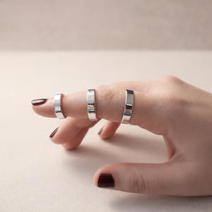 May include: Three silver rings are worn on the fingers of a hand. The rings are flat and rectangular in shape. The background is a solid, neutral color.