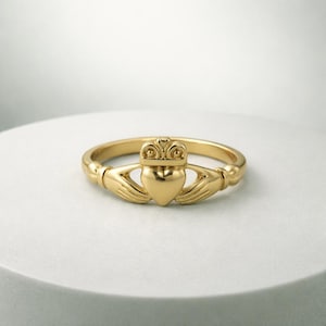 May include: A gold Claddagh ring, a traditional Irish design. The ring features a heart, hands, and a crown. The heart is in the center, held by two hands, and topped with a crown. The ring is displayed on a white surface.