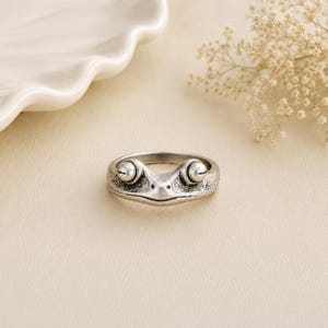 May include: A silver-toned metal ring featuring a sculpted frog design. The band displays a textured, weathered finish with two prominent, rounded eyes protruding from the top and a small, pointed snout centred between them.